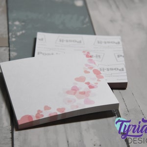 May include: A white sticky note pad with a pink and white watercolour design of hearts. The pad is on a grey wooden surface. The text "Post-it" is visible on the back of the pad.