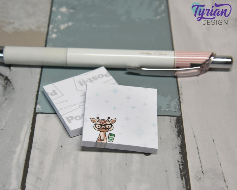 May include: A white notepad with a blue and white starry pattern. A cartoon giraffe wearing glasses and holding a cup of coffee is printed on the notepad. The notepad is on a wooden surface.