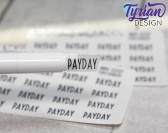 May include: A white marker with a black sticker that says "PAYDAY" on it. The sticker is slightly curled at the edges. The marker is on top of a sheet of paper with the word "PAYDAY" printed on it multiple times.
