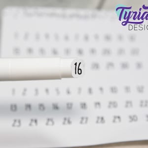 May include: A white marker with a purple cap and a white sticker with the number 16 on it. The marker is in focus and the sticker is blurred. The background is a calendar with the numbers 1 through 31.
