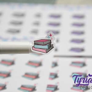 May include: A stack of three books with a pink, blue, and white cover. The books are decorated with a simple line drawing style. There are two small stars above the books.
