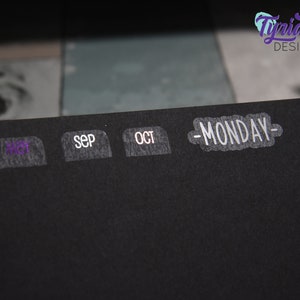 K&ouml;nnte beinhalten: Ein schwarzer Planer mit wei&szlig;en Etiketten f&uuml;r die Monate August, September, Oktober und einem wei&szlig;en Etikett mit dem Text "MONDAY" darauf.