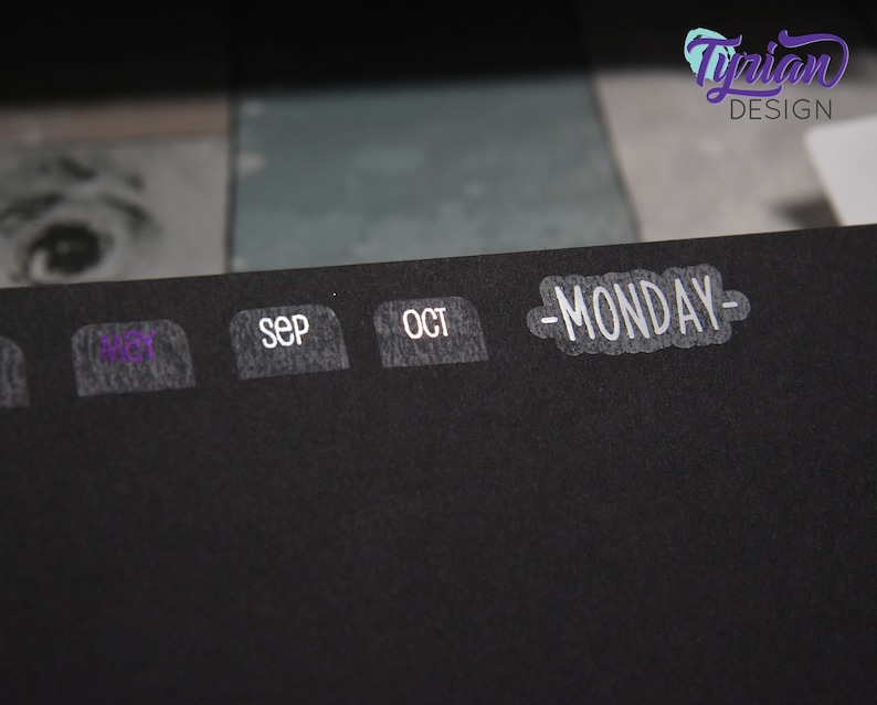 K&ouml;nnte beinhalten: Ein schwarzer Planer mit wei&szlig;en Etiketten f&uuml;r die Monate August, September, Oktober und einem wei&szlig;en Etikett mit dem Text "MONDAY" darauf.