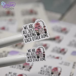 May include: Two white stickers featuring the black text WE ARE ALL JUST WALKING EACH OTHER HOME, accompanied by a graphic of a white picket fence, a garden arch, and pink roses. The stickers are displayed on white backing paper.