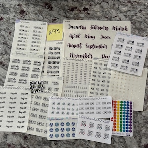 May include: A collection of planner stickers on a speckled surface. The stickers include labels for holidays, months, days of the week, and various icons. Some stickers have text like "Happy Birthday" and "Dream Log". There are also colorful dot stickers.