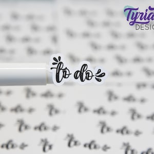 May include: A close-up of a black and white "to do" sticker with a small flower design. The sticker is on top of a white pen.