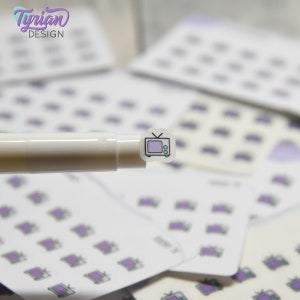May include: A close-up of a clear sticker with a purple and black outline of a vintage television set. The sticker is on a white pen.