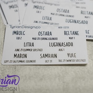 May include: A white card with the names of the eight pagan holidays, including Imbolc, Ostara, Beltane, Litha, Lughnasadh, Mabon, Samhain, and Yule, with their corresponding dates and descriptions.