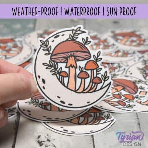 May include: A sticker featuring a crescent moon with a cluster of mushrooms in shades of brown and orange. The mushrooms are surrounded by green leaves and the sticker has a white background.