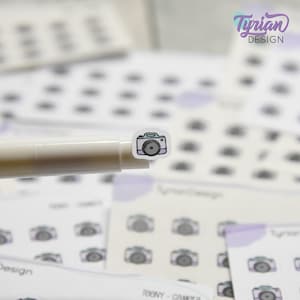 May include: A sheet of stickers featuring a cartoon camera design. The stickers are white with a light purple and teal outline. The camera is a light teal colour with a black lens.