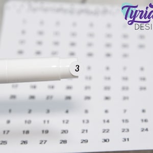 May include: A white marker with a purple stripe and a white circle sticker with the number "3" on it. The marker is on top of a calendar with the numbers 10 through 31 visible.