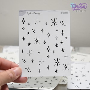 May include: A sheet of white stickers featuring a pattern of black stars and starbursts. The text "TyrianDesign" and "ST-10542" are printed on the sticker sheet. The stickers are held by a hand.