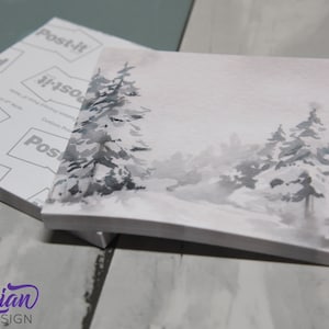 May include: A pack of Post-it Notes featuring a watercolour design of a snowy woodland scene. The design depicts grey and white trees against a light grey background.