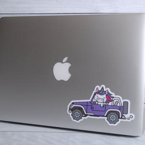 Puede incluir: Una pegatina de un jeep morado con un unicornio conduciendo. El unicornio tiene una crin y una cola azul, rosa y blanca. El jeep tiene ruedas negras y un fondo blanco.
