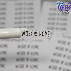 May include: A close-up of a white sticker with the text "WORK @ HOME" in black, held by a white pen. The sticker is surrounded by other stickers with the same text, all on a white background. The logo "Tyrian Design" is visible in the upper right corner.