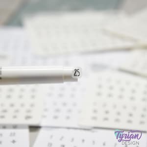 May include: A white pen with the number 25 printed on a small, clear sticker. The pen is in focus, with a blurred background of white paper with printed numbers. The image includes the text "Tyrian Design".