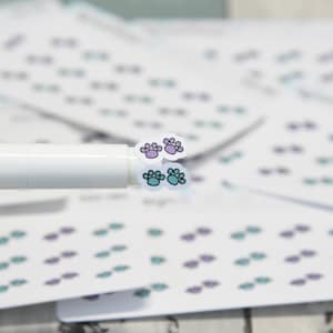 May include: A sheet of stickers with paw prints in teal and purple. The stickers are on a white background and are arranged in rows. A white marker is holding two of the stickers.