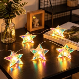 May include: A string of star-shaped LED lights featuring a faceted, iridescent finish that reflects multiple colors. Each star contains a warm glowing light, connected by a thin, flexible wire, creating a luminous display of geometric shapes.