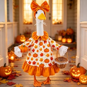 May include: A white goose figurine wearing an orange bow headband, a ruffled collar, and a white dress patterned with orange pumpkins and autumn leaves. The goose holds a small woven basket in one wing and a miniature orange pumpkin in the other.