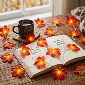 May include: A string of LED fairy lights featuring artificial maple leaves in shades of red, orange, and yellow. The lights are draped over an open book titled Autumn Harvest Stories and a dark ceramic mug resting on a gray knitted coaster.