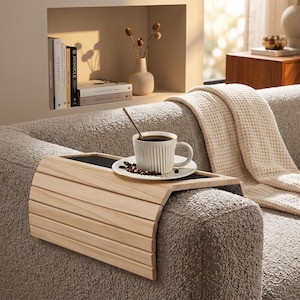 May include: A flexible wooden armrest tray with a light natural finish, designed to drape over a sofa arm. The tray features a flat, dark central surface that supports a white ribbed ceramic mug filled with coffee and a small plate of coffee beans.