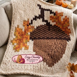 Pode incluir: Uma manta de malha grossa na cor creme com um design de uma grande bolota castanha e folhas de outono. Um logótipo oval está impresso no canto inferior esquerdo com o texto Cozy Chunky BLANKETS by Sharon e a imagem de uma pessoa.