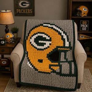 May include: A chunky knit throw blanket featuring a large, centered graphic of a yellow, white, and dark green football helmet. The blanket has a thick, textured yarn construction with a dark green border framing the grey background.