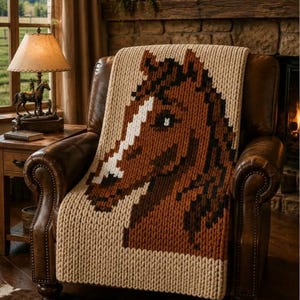 May include: A chunky knit throw blanket featuring a large, pixelated horse head design in shades of brown, dark brown, and white, set against a light beige background. The blanket is draped over a brown leather armchair, showcasing its thick texture.