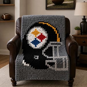May include: A chunky knit throw blanket featuring a large pixelated graphic of a black football helmet. The helmet design includes a white circle with yellow, red, and blue star shapes, a yellow stripe, and a gray face mask, all on a gray background.
