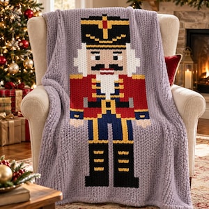 Könnte beinhalten: Eine grob gestrickte Wohndecke in hellem Lavendel mit einem großen, pixeligen Nussknacker-Motiv in der Mitte. Der Nussknacker ist mit einem schwarzen Hut, einer roten Jacke, einer blauen Hose und schwarzen Stiefeln in einem blockartigen Rastermuster dargestellt.