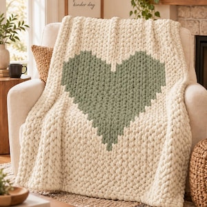Könnte beinhalten: Eine grob gestrickte Wohndecke mit einem großen, salbeigrünen Herzmuster, das auf einem cremefarbenen Hintergrund zentriert ist. Die Decke ist aus dickem, weichem Garn mit einer markanten Zopfstrickstruktur gefertigt, was ihr ein gemütliches Aussehen verleiht.