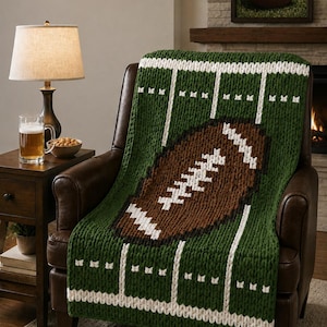 May include: A chunky knit throw blanket featuring a green football field design with white yard lines and a large brown football in the center. The blanket is crafted with thick yarn, showcasing a textured pattern that mimics a sports field layout.
