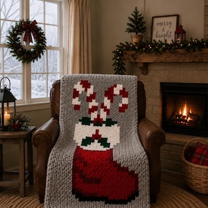 Könnte beinhalten: Eine grob gestrickte Wohndecke in Hellgrau mit einem großen, pixeligen Motiv eines roten Weihnachtsstrumpfes, gefüllt mit zwei Zuckerstangen, weißem Rand und grünen Stechpalmenblättern, drapiert über einem braunen Ledersessel.