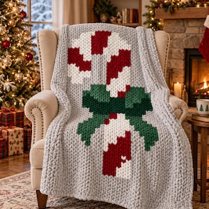 May include: A chunky knit throw blanket in light grey featuring a large, centred candy cane design with red and white stripes, accented by a dark green bow. The blanket is draped over a beige upholstered armchair with visible fabric texture.