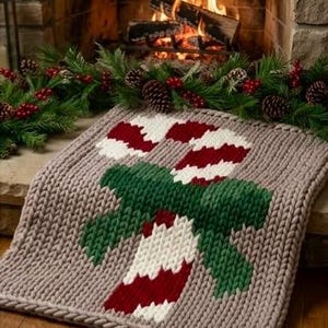 May include: A chunky knit blanket in a light taupe color featuring a large, centered candy cane design in red and white, accented by a dark green bow. The blanket is crafted with thick, visible yarn stitches, creating a textured, soft appearance.