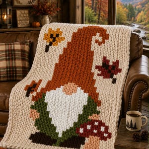 May include: A chunky knit throw blanket in cream, featuring a large gnome graphic with a tall orange hat, a white beard, green clothing, and a red mushroom. The design includes scattered autumn leaves in yellow and dark red colors.