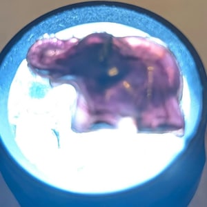 Könnte beinhalten: Eine kleine, durchscheinende violette Elefantenfigur wird von einer hellen, kreisförmigen Lichtquelle von hinten beleuchtet. Der Elefant ist im Profil dargestellt, blickt nach links mit leicht erhobenem Rüssel, wobei das Licht durchscheint.
