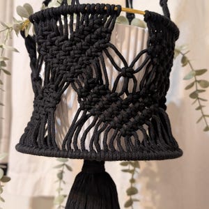 Peut inclure: Une suspension pour plante en macramé noir avec des motifs noués complexes et un pompon central. Elle soutient un pot en céramique blanche cannelée avec un bord en métal doré, contenant une plante verte retombante aux petites feuilles.