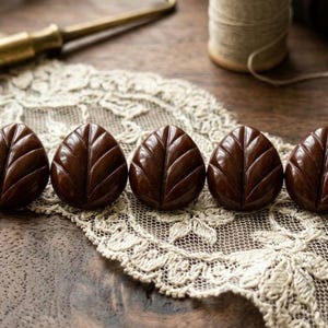 May include: Five dark brown buttons, each shaped like a rounded leaf with a central vein and symmetrical side veins carved into the surface. The buttons have a smooth, glossy finish and are arranged in a horizontal row on a piece of lace.