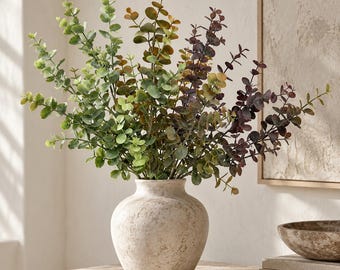 May include: A round, beige, textured ceramic vase holds a large arrangement of artificial eucalyptus stems. The foliage displays a gradient of colors, transitioning from bright green on the left to deep reddish-brown on the right side.