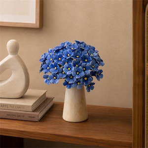 Può includere: Un bouquet in ceramica composto da fiori blu vivaci con piccoli centri gialli, disposto in un vaso affusolato color bianco sporco. La scultura presenta numerosi fiori singoli con petali testurizzati, creando una composizione densa.