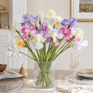 May include: A bouquet of artificial iris flowers in shades of white, yellow, pink, and purple arranged in a clear, vertically ribbed cylindrical glass vase. The flowers feature ruffled petals and green stems, creating a vibrant floral display.
