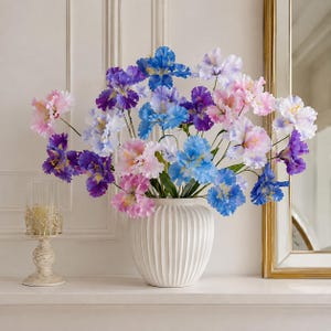 Può includere: Un bouquet di fiori di iris artificiali nelle tonalità del blu, viola e rosa, disposto in un vaso di ceramica bianco a coste verticali. I fiori presentano petali delicati e increspati con centri gialli e steli verdi, creando un display vivace.