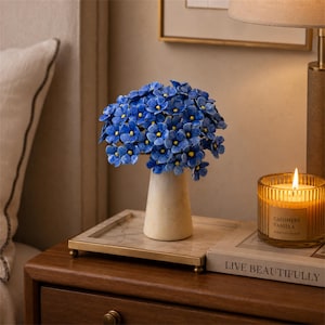 May include: A ceramic vase filled with a dense cluster of small, vibrant blue flowers featuring bright yellow centres. The vase has a smooth, tapered, off-white body and sits on a rectangular tray with a metallic gold-toned frame and base.
