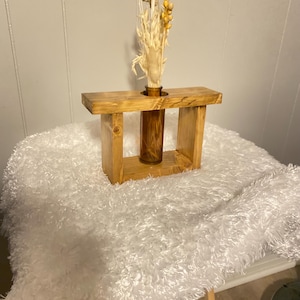 May include: A rectangular wooden frame with a natural brown finish, featuring a central amber-colored glass bud vase. The vase holds a collection of dried beige and light yellow botanical stems, including grasses and small seed pods.
