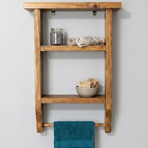 May include: A wall-mounted wooden shelf unit with a medium brown finish, featuring two horizontal shelves and a bottom towel bar. The top shelf holds a glass jar and rolled fabric, while the lower shelf holds a bowl with soap bars.