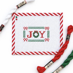 May include: A rectangular needlepoint canvas featuring the word JOY stitched in red and green, framed by a red and white candy cane striped border. Three skeins of embroidery floss in white, red, and green are positioned around the canvas.