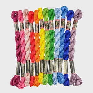 May include: A collection of twelve twisted skeins of DMC Coton Perlé embroidery thread arranged in a spectrum of colors including magenta, pink, light pink, red, orange, yellow, lime green, green, light blue, blue, dark blue, and lavender.