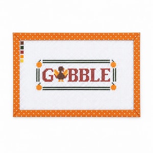 May include: A rectangular cross-stitch pattern on white fabric featuring the text GOBBLE in red letters, with a brown turkey replacing the letter O. The design is framed by an orange border with white polka dots and includes a colour key.