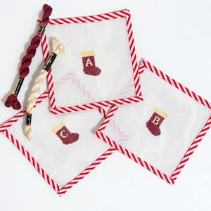 May include: Three square needlepoint canvases with red and white striped fabric borders. Each canvas features a small stitched maroon Christmas stocking with a yellow cuff, labeled with the letters A, B, and C. Two skeins of embroidery floss are included.